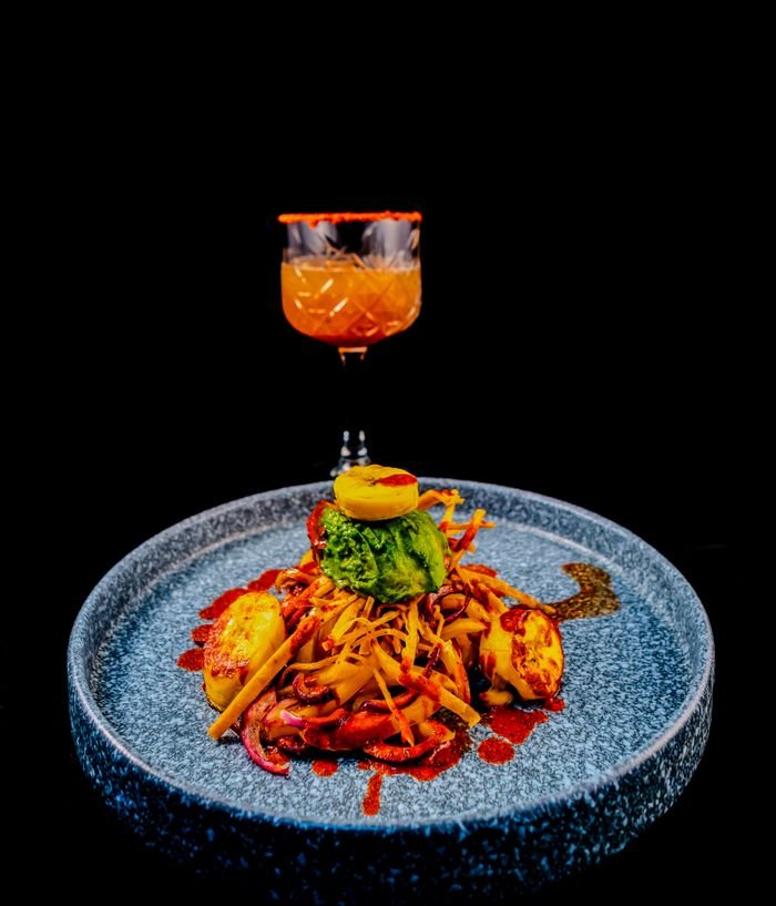 A vibrant gourmet dish paired with a cocktail, showcasing elegant culinary artistry against a black background.
